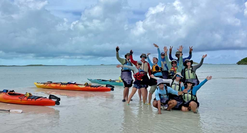 A group of people pose for a group photo in ankle deep water. Many of them raise their hands in celebration. There are several empty kayaks beside them. 